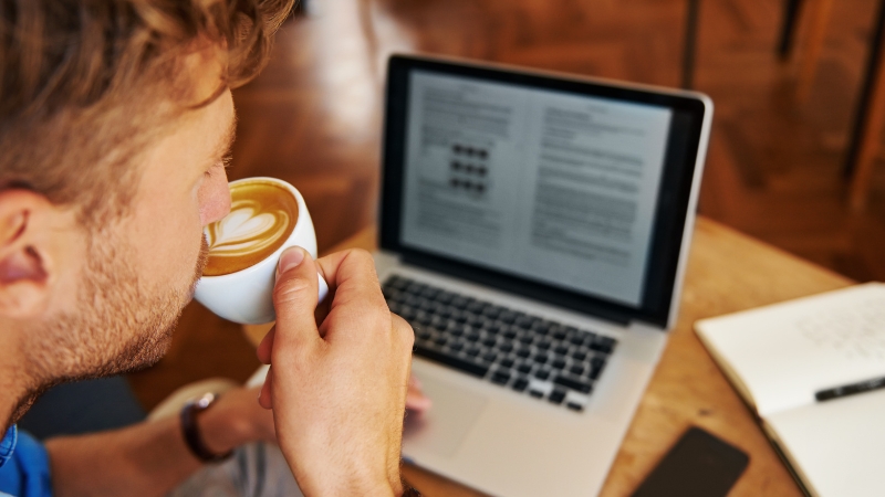 Person drinking coffee while reviewing an article draft on a laptop at a work desk
